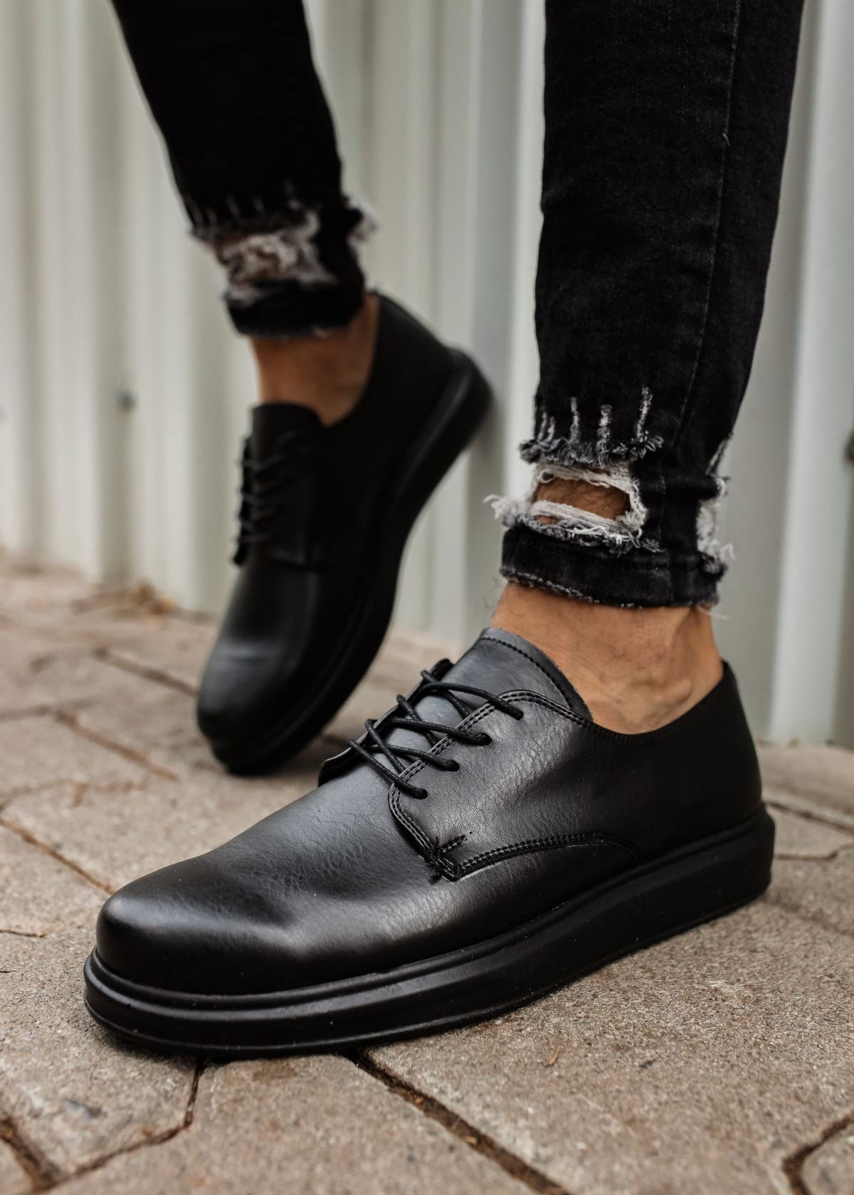 A pair of men's black dress shoes with lace-up closure, worn by a person against an outdoor background.